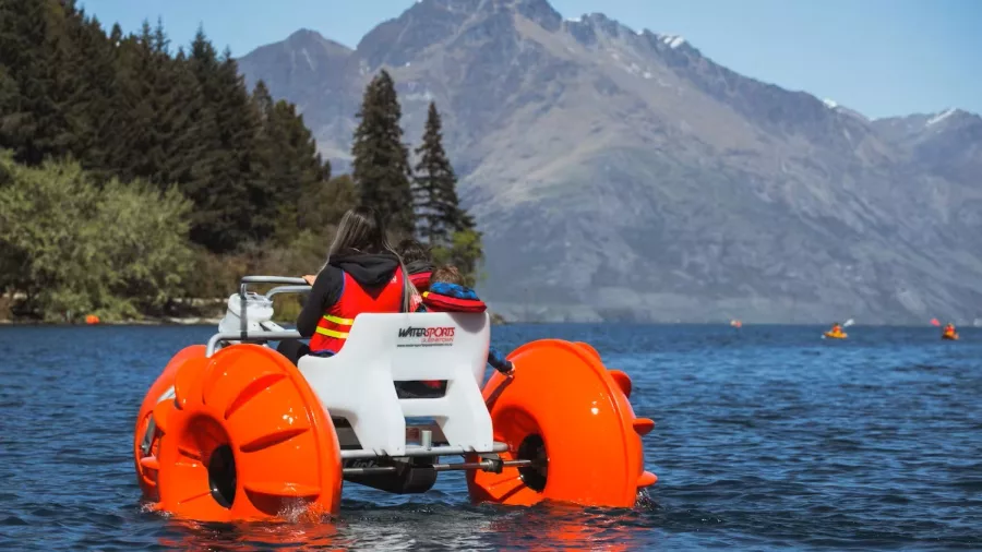 Person riding a bright orange aqua bike on Lake Whakatipu with mountains in background
