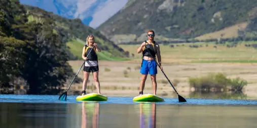 Two people stand-up paddleboarding on calm water surrounded by Queenstown mountains.