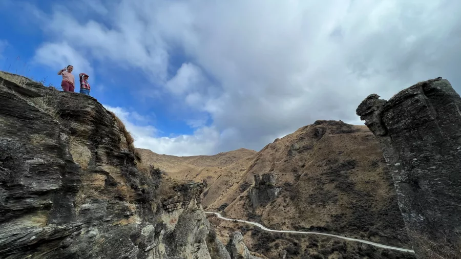 Two people standing on a high rock ledge overlooking the winding road of Skippers Canyon