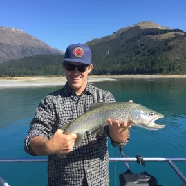 Man holding a trout on a fishing boat with mountains behind Lake Wakatipu