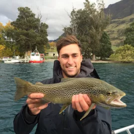 Young man holding a brown trout beside a fishing boat on Lake Wakatipu