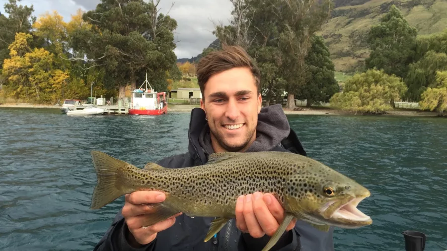 Young man holding a brown trout beside a fishing boat on Lake Wakatipu