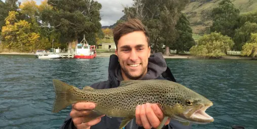 Young man holding a brown trout beside a fishing boat on Lake Wakatipu
