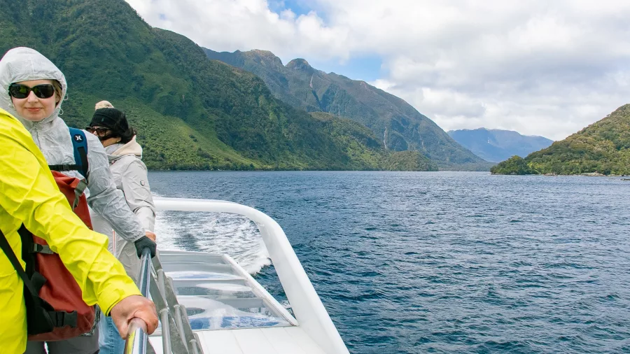 Tourists wearing jackets on the deck of a boat cruising through Patea Arm in Doubtful Sound, surrounded by lush mountains