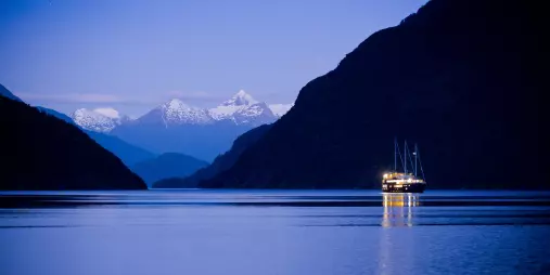 Fiordland Navigator lit up at dusk with snow-covered peaks in the distance