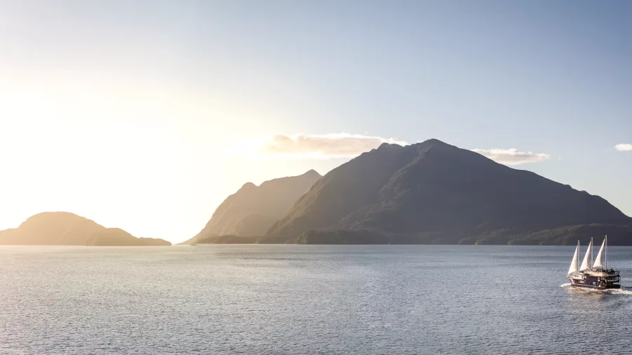 Fiordland Navigator with sails raised under a glowing sunset in Doubtful Sound with silhouetted mountains in the distance