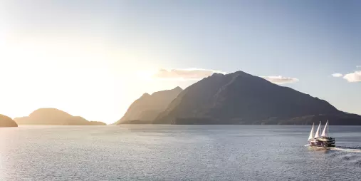 Fiordland Navigator with sails raised under a glowing sunset in Doubtful Sound with silhouetted mountains in the distance