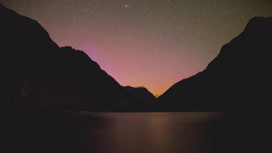 Starry night sky over Milford Sound with silhouetted mountains and soft light glow on the horizon