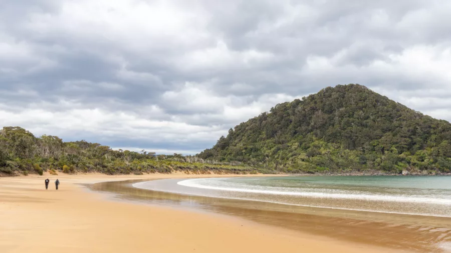 Two people walking along a vast, deserted beach beside forested hills in southern Fiordland