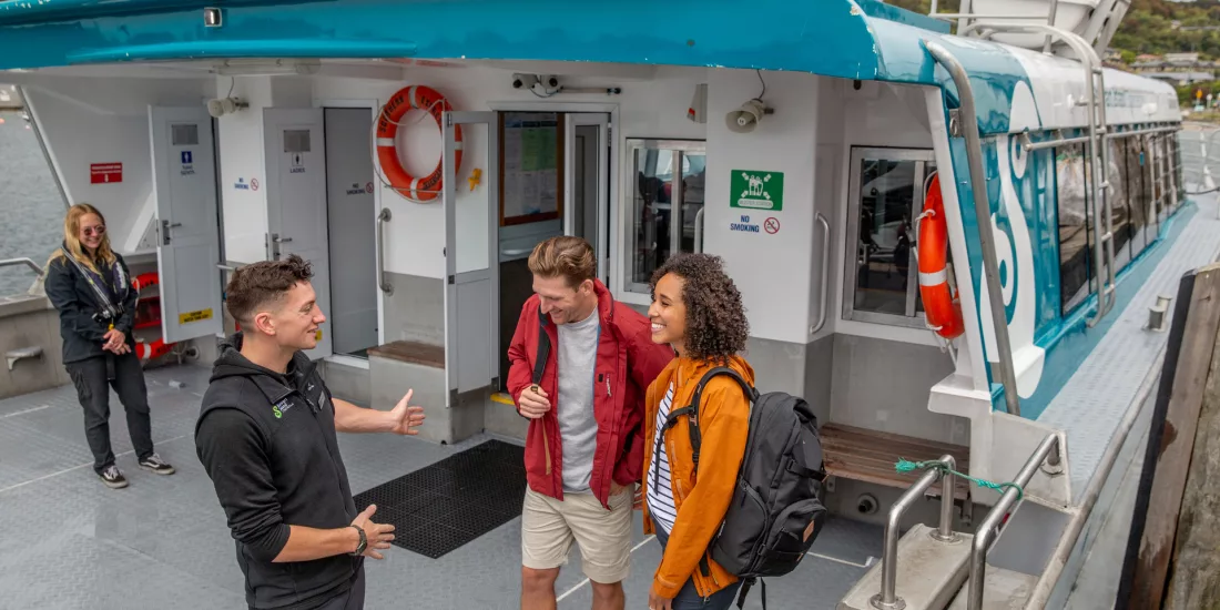 A ferry crew member greeting two passengers aboard the boat, with the Stewart Island ferry terminal in the background.