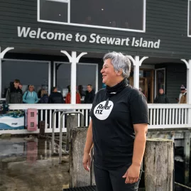A staff member at the Stewart Island Ferry terminal welcomes visitors with a "Welcome to Stewart Island" sign in the background.