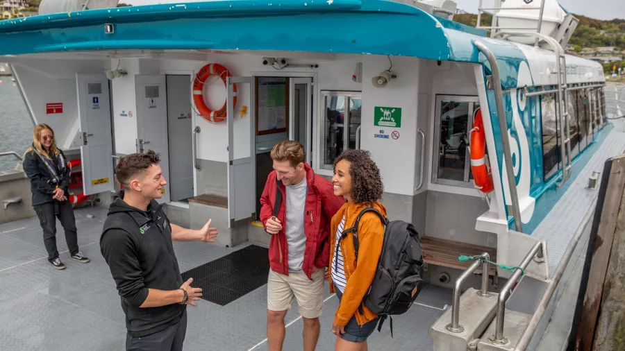 A ferry crew member greeting two passengers aboard the boat, with the Stewart Island ferry terminal in the background.