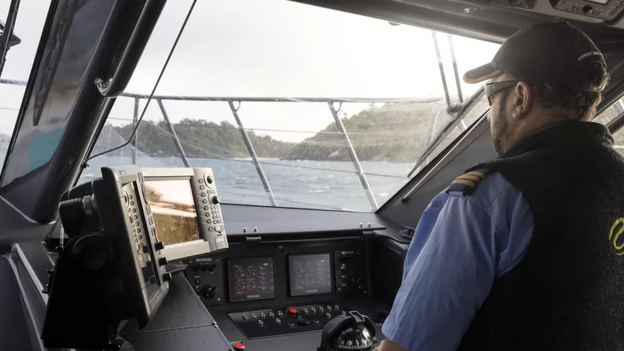 Captain steering the ferry, with the control panel and coastal view visible from the bridge.