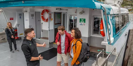 A ferry crew member greeting two passengers aboard the boat, with the Stewart Island ferry terminal in the background.