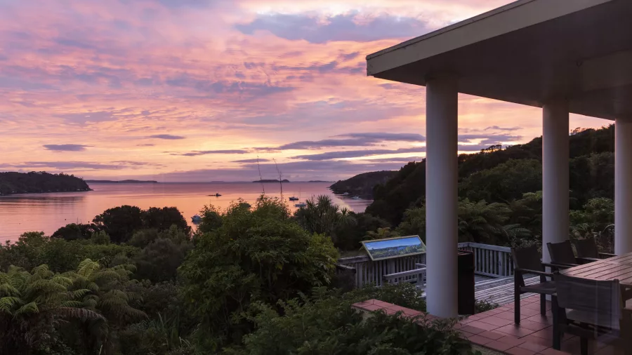 Vibrant sunset over a coastal area on Rakiura Stewart Island, with boats on calm water and a patio area featuring columns, a table, and chairs in the foreground.