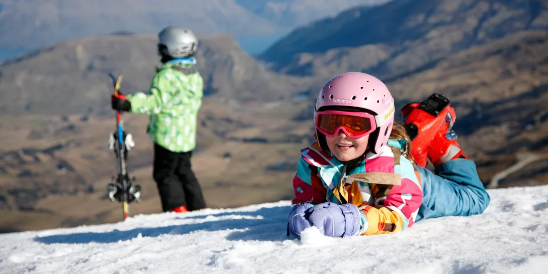 Young children enjoying the snow at Coronet Peak ski field, with alpine views over Queenstown in the background