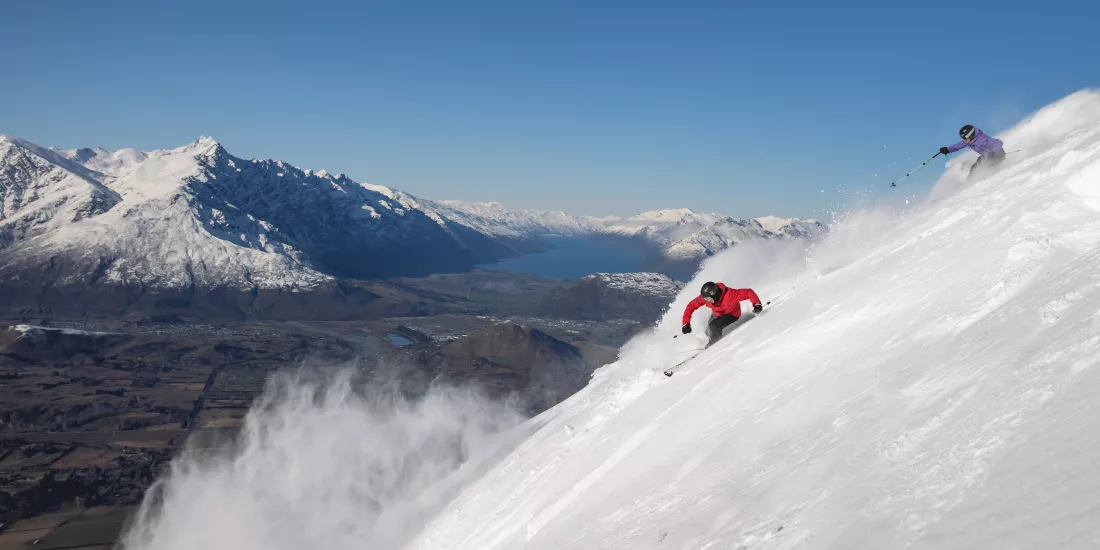 Two skiers carving through powder snow at Coronet Peak with a panoramic view of Lake Wakatipu and the Remarkables