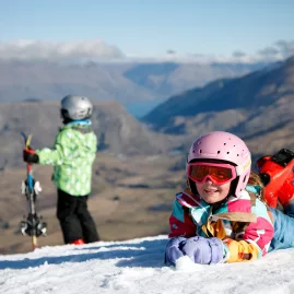 Young children enjoying the snow at Coronet Peak ski field, with alpine views over Queenstown in the background