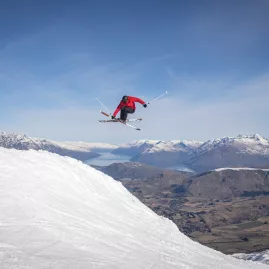 Freestyle skier catching air off a jump at Coronet Peak with snowy mountains and Lake Wakatipu in the distance