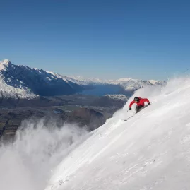 Two skiers carving through powder snow at Coronet Peak with a panoramic view of Lake Wakatipu and the Remarkables