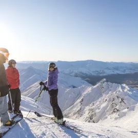 Three skiers standing on a snowy ridge at Coronet Peak with panoramic views over Queenstown and surrounding mountains