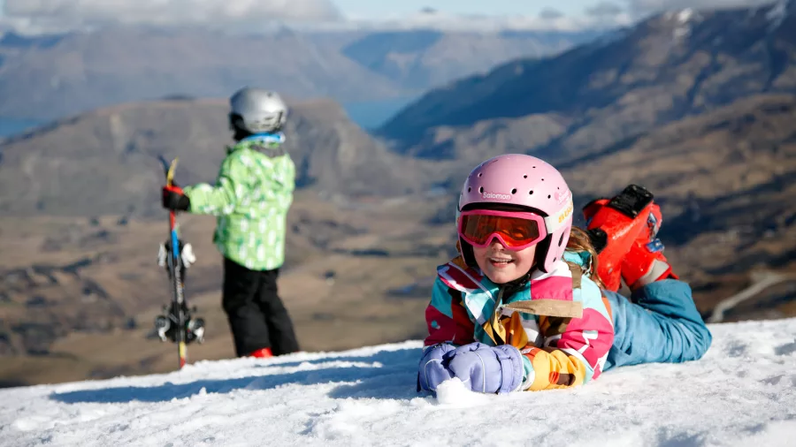 Young children enjoying the snow at Coronet Peak ski field, with alpine views over Queenstown in the background