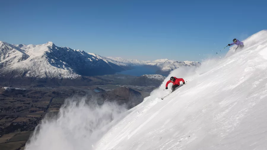 Two skiers carving through powder snow at Coronet Peak with a panoramic view of Lake Wakatipu and the Remarkables