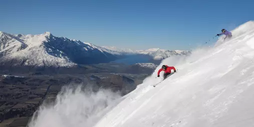 Two skiers carving through powder snow at Coronet Peak with a panoramic view of Lake Wakatipu and the Remarkables