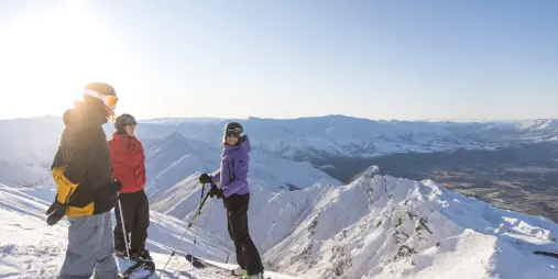 Three skiers standing on a snowy ridge at Coronet Peak with panoramic views over Queenstown and surrounding mountains