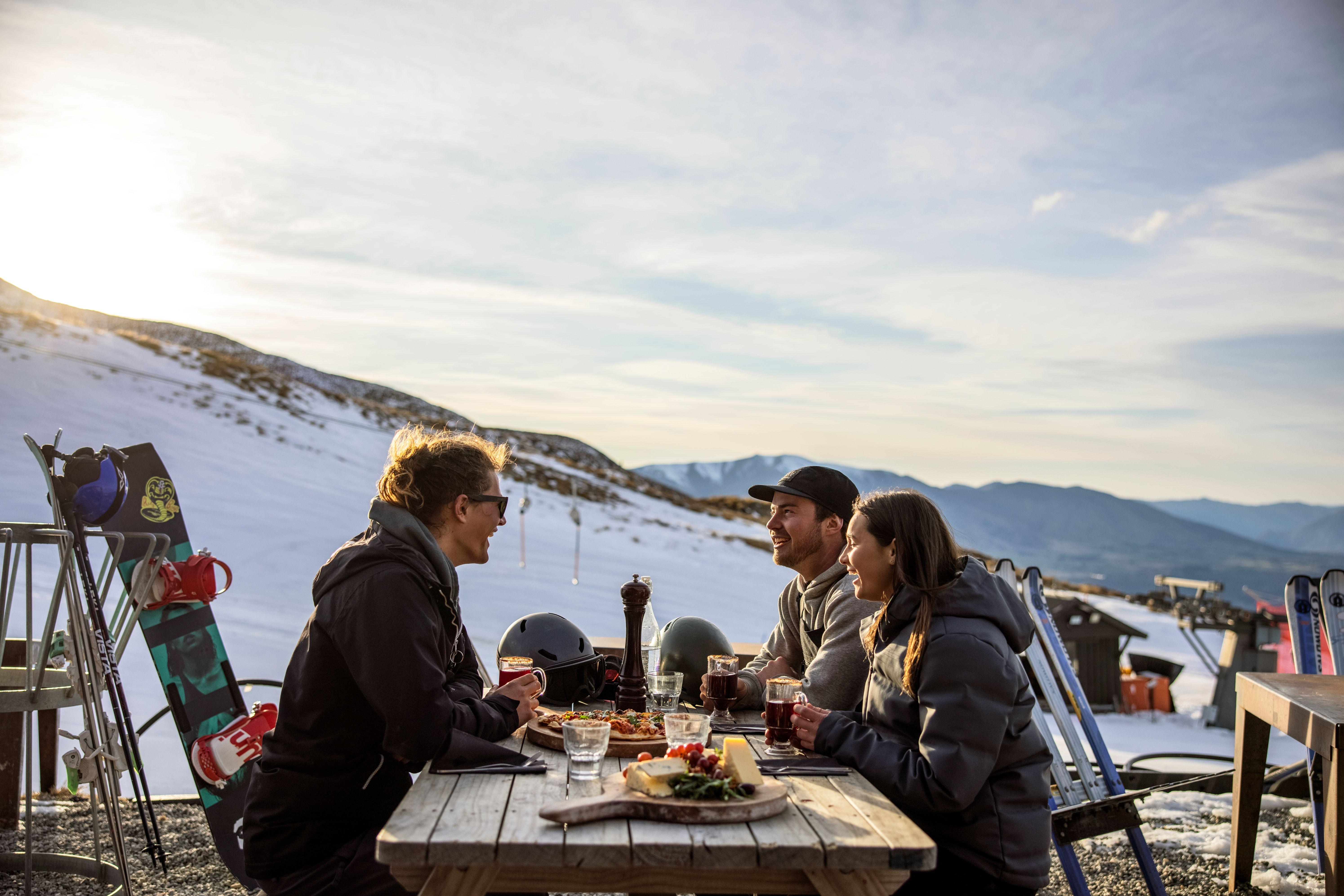 Friends enjoying après ski meal outdoors at Coronet Peak with snow and mountain backdrop