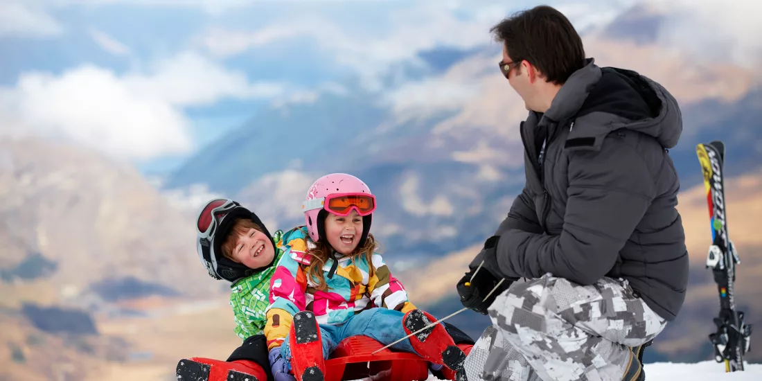 Two young children with a parent sitting on the snow at The Remarkables ski resort in Queenstown
