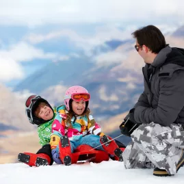 Two young children with a parent sitting on the snow at The Remarkables ski resort in Queenstown