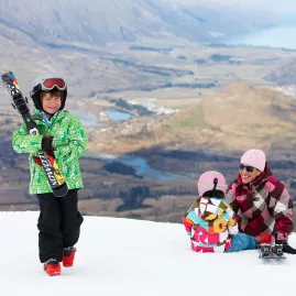 Child carrying skis while family sits on the slope behind at The Remarkables ski resort in Queenstown