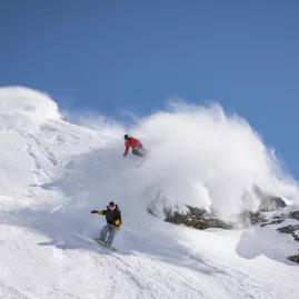 Three riders skiing and snowboarding through fresh powder on a steep slope at The Remarkables