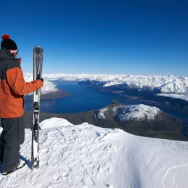 Skier holding skis and looking out over Lake Wakatipu from a snowy ridge at The Remarkables ski field