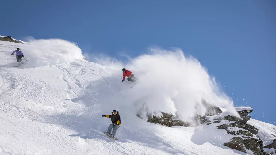 Three riders skiing and snowboarding through fresh powder on a steep slope at The Remarkables