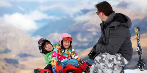 Two young children with a parent sitting on the snow at The Remarkables ski resort in Queenstown