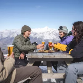 Friends enjoying drinks at an outdoor table at Heidi's Hut with snowy mountain views at Coronet Peak, Queenstown
