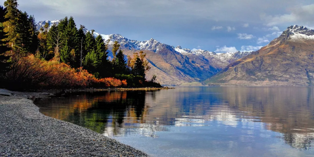 Peaceful shoreline of Lake Wakatipu with autumn trees and snow-capped mountains in Queenstown