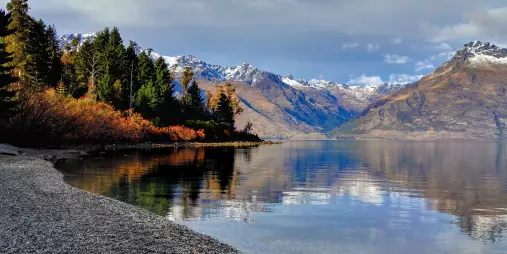 Peaceful shoreline of Lake Wakatipu with autumn trees and snow-capped mountains in Queenstown