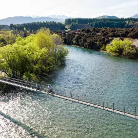 Cyclists crossing the Hāwea River swing bridge on a sunny day in Wanaka