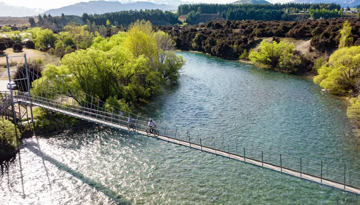 Cyclists crossing the Hāwea River swing bridge on a sunny day in Wanaka