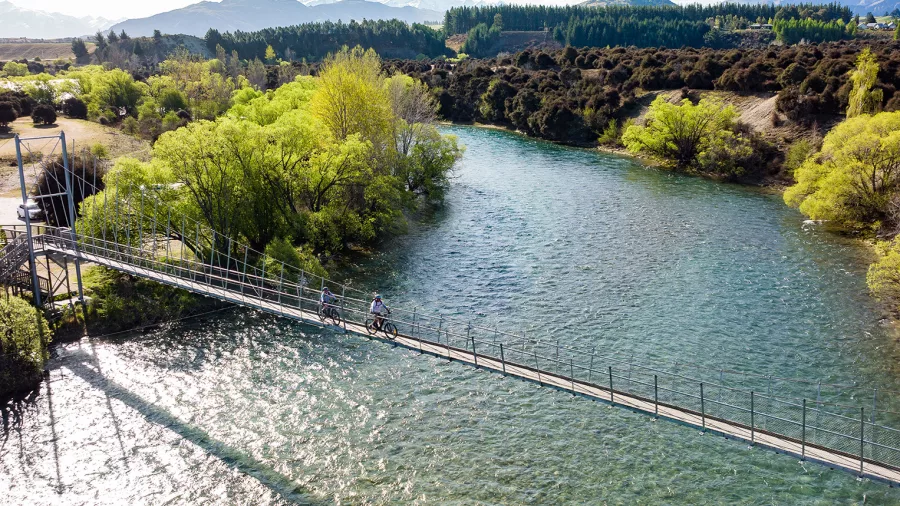 Cyclists crossing the Hāwea River swing bridge on a sunny day in Wanaka