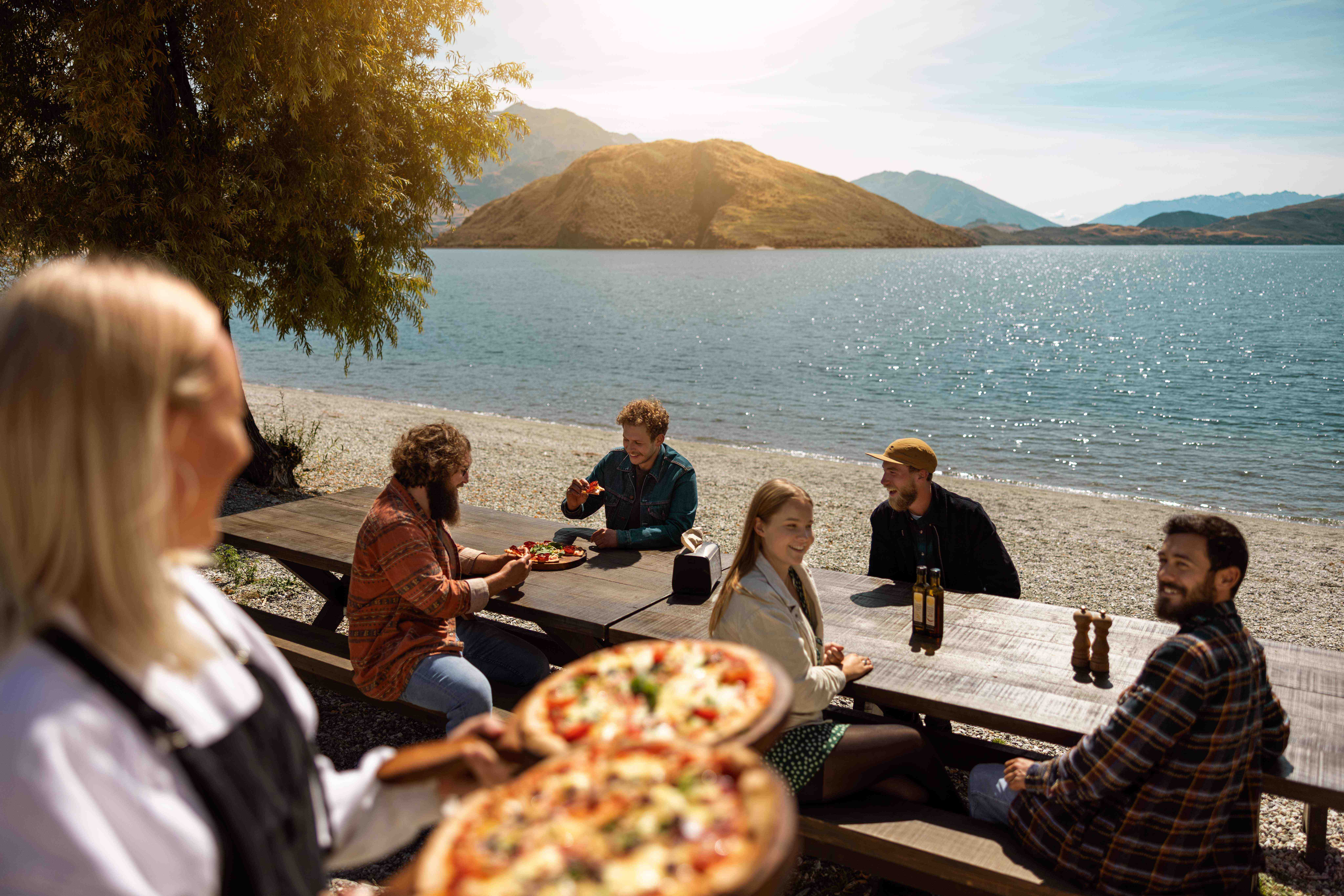 A group of people eating pizza at picnic tables beside Lake Wānaka in Otago, New Zealand, with the lake, mountains, and bright afternoon sun in the background.