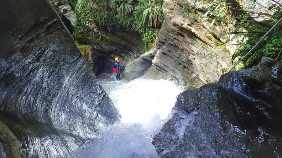 Canyoner descending through a narrow rock chute above a deep pool in a forest canyon near Wanaka.