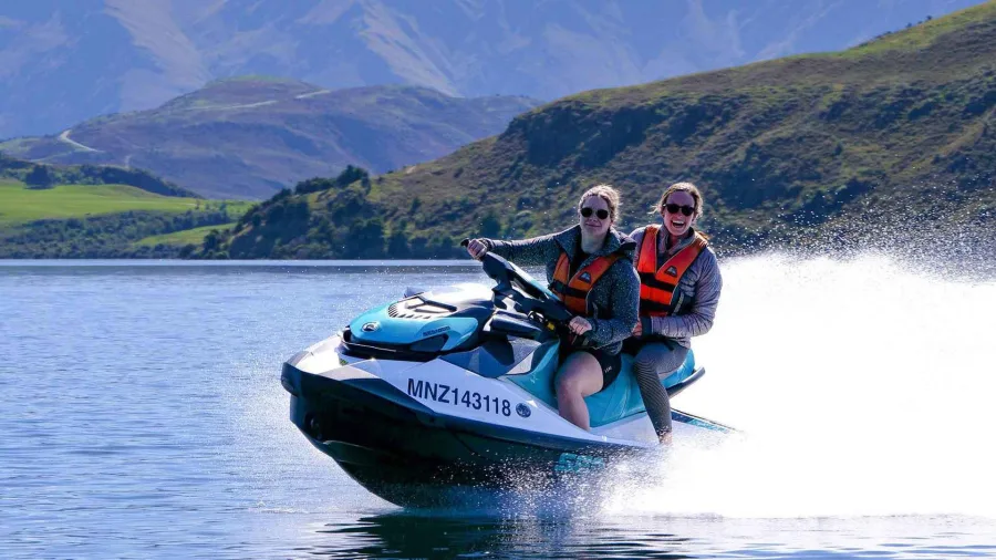 Two smiling women riding a jet ski on Lake Wanaka with scenic hills in the background.