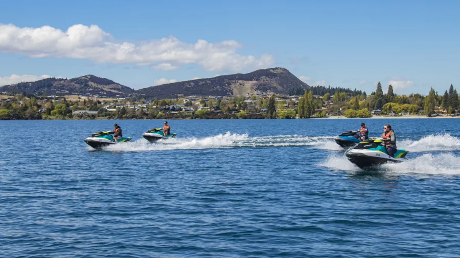 Group of four jet skis speeding across Lake Wanaka with Wanaka township and mountains in the background.