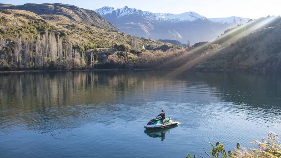 Solo rider on a jet ski in a quiet cove on Lake Wanaka with sunbeams and snow-capped mountains.