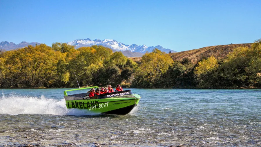 Jet boat speeding along the Clutha River with passengers on board and snow-capped mountains in the background.