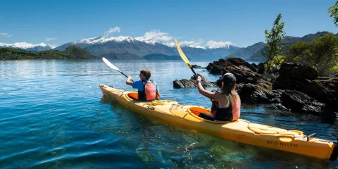 Two people paddling a double kayak near the shore of Lake Wanaka with mountain views.